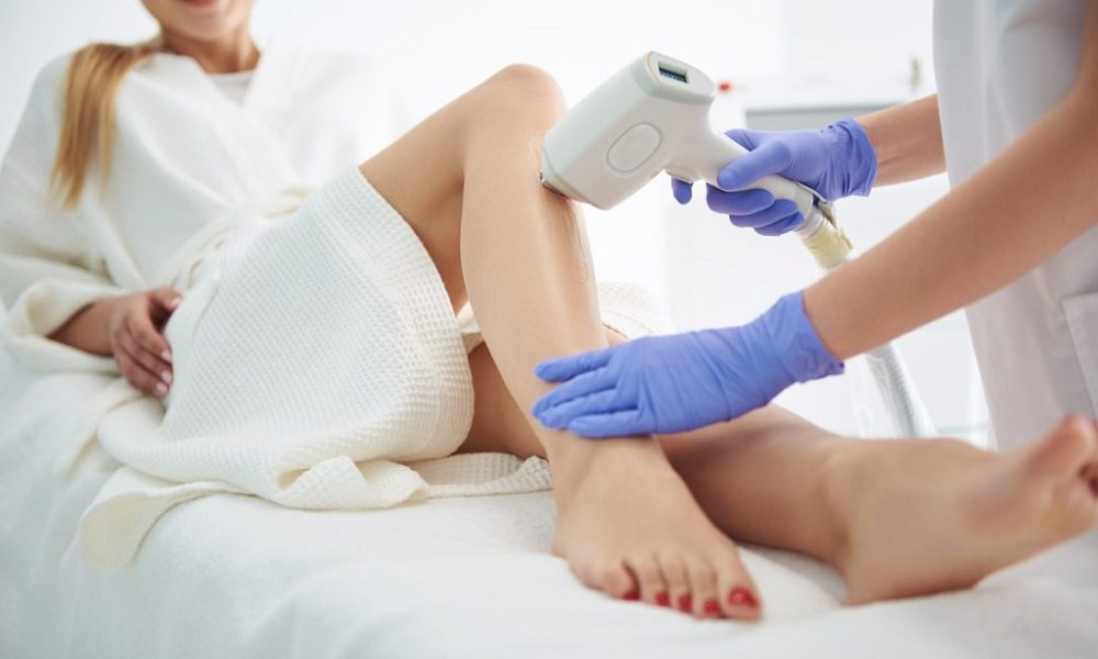 Close up of beautician in sterile blue gloves placing laser epilation machine on lady leg with clear gel and gliding device over the skin. Lady lying on daybed and smiling